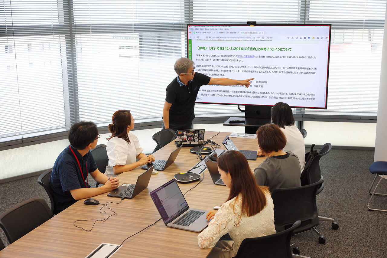 大きなモニター画面を指差しながら説明している植木氏と話に聞き入るプロジェクトメンバーたち