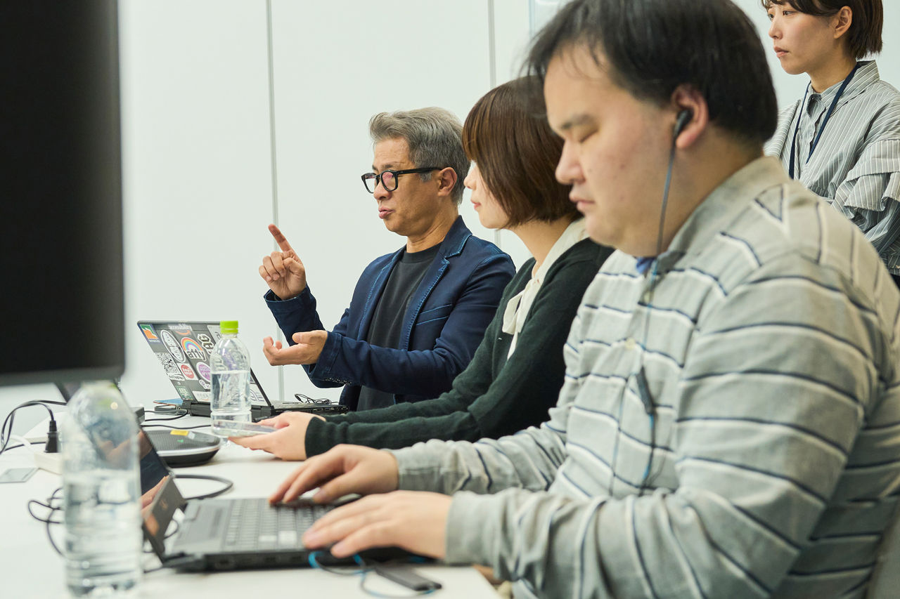 写真：スクリーンリーダーの音声をイヤホンで聞きながら操作している北村さんと、奥の座席で補足説明をしている植木さん