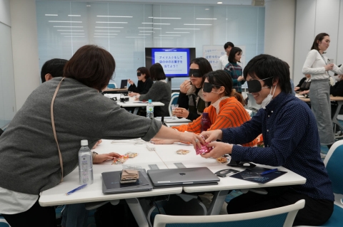 写真：5名の参加者がアイマスクをしてテーブルを囲み、お菓子を手渡ししている