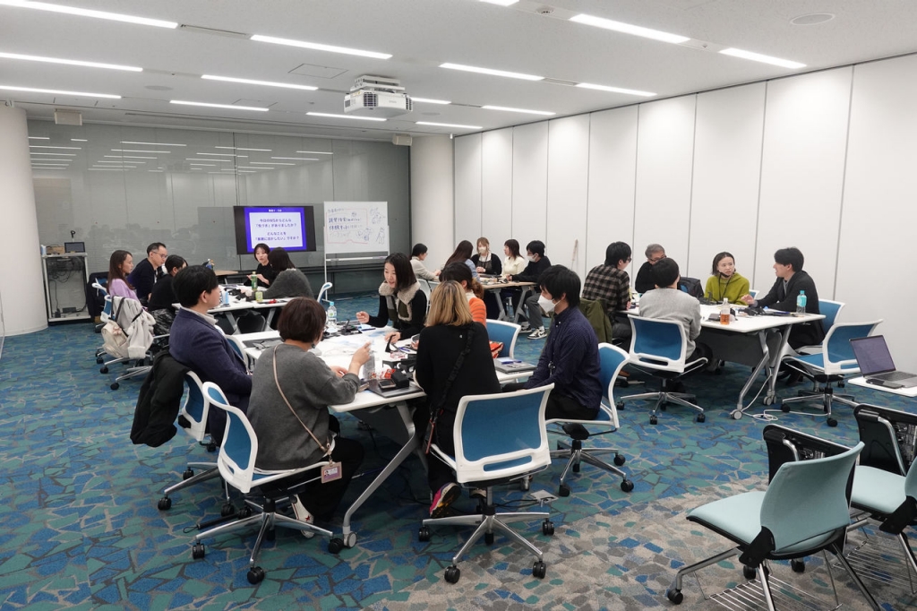 写真：写真：5～6名ずつで4つのグループに分かれて、それぞれテーブルを囲んでいる参加者