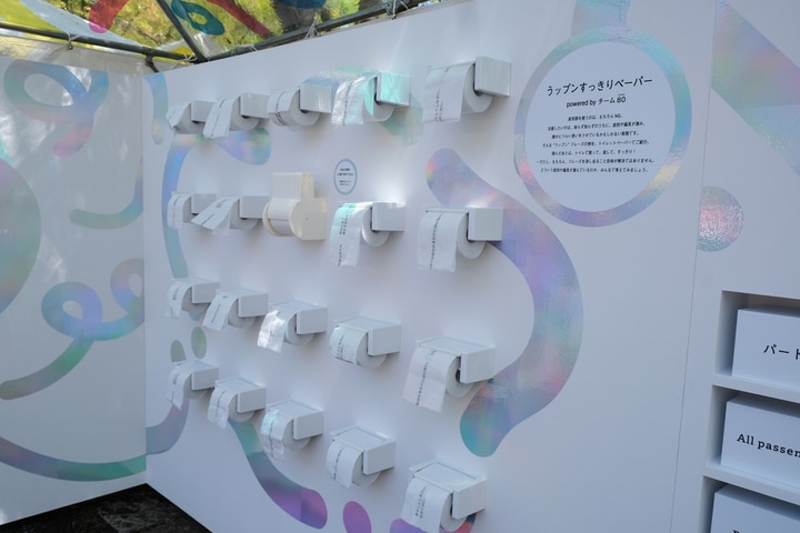 うップンすっきりペーパーの展示の様子を写したもの。高さ2メートルほどのブースの白を基調とした壁一面に、5列かける4段、合計20個のトイレットペーパーホルダーが取り付けられている。ホルダーにはそれぞれうップンすっきりペーパーがセットされている。