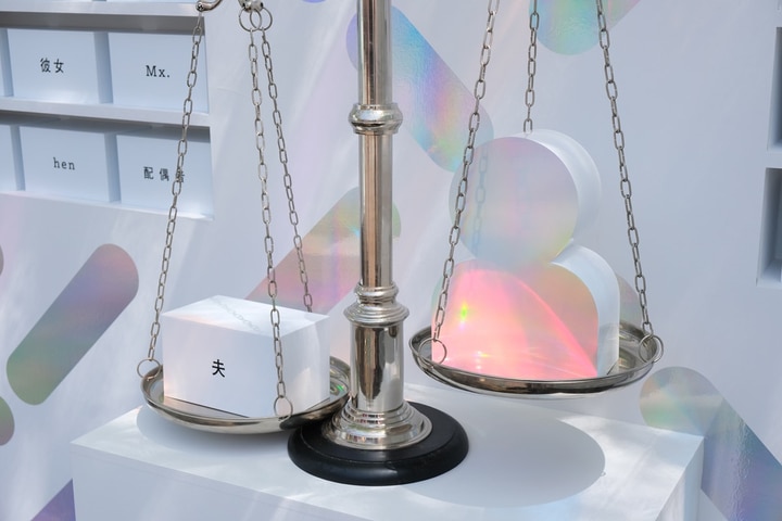 言葉の天秤の展示の様子。白を基調とした壁に設けられた3段の棚に、さまざまな言葉が書かれた白いブロックが収められている。書かれている言葉は、「Mr.」「she」「●●さん」「Ladies and Gentlemen」「（ひらがなで）きょうだい」「hen」「夫」「恋人」「Mx.」など