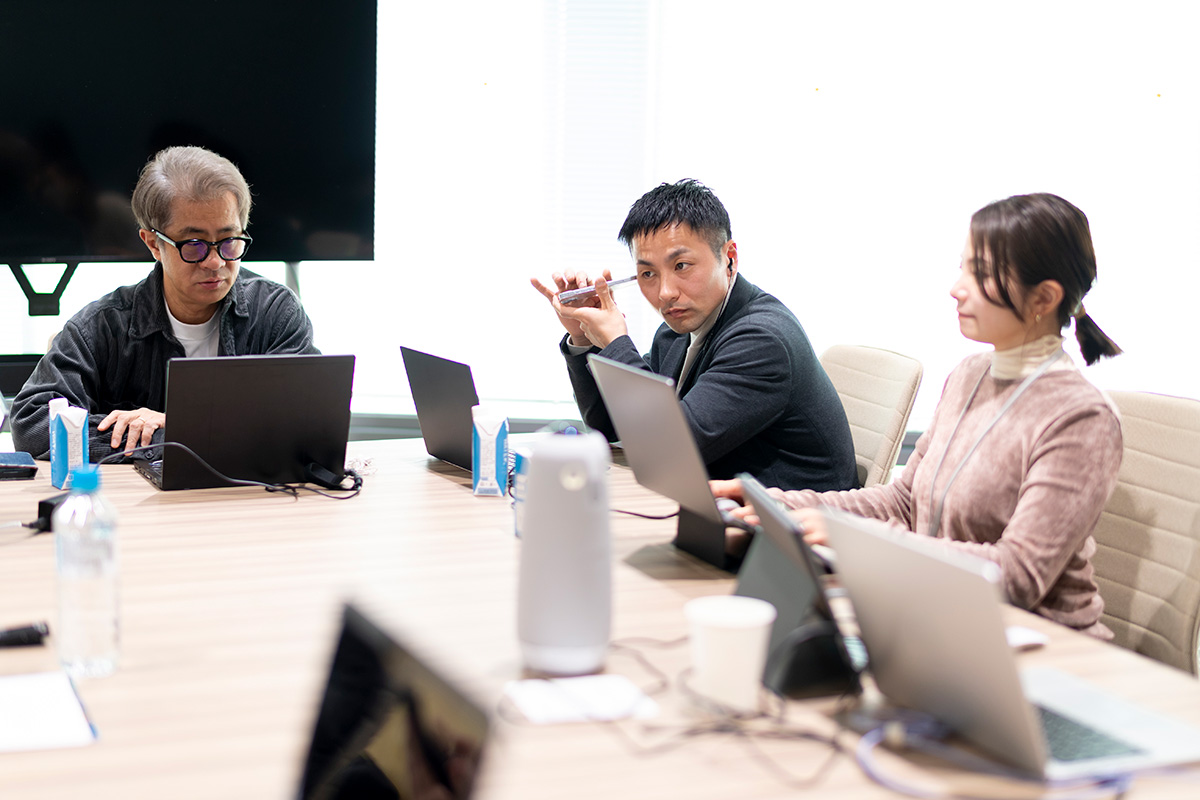 写真：スマートフォンを耳に近づけて、音声を聞く川嶋選手と、それを見守るアクセシビリティプロジェクトメンバー2名